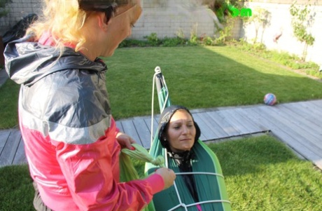 Blonde girl encases her girlfriend in rainwear on a hammock before soaking her 26654125
